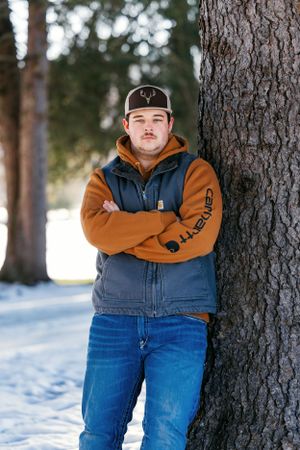 Person wearing baseball cap and brown Carhartt jacket poses against tree in winter setting.