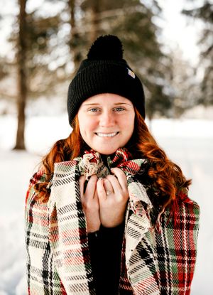 Winter portrait with plaid blanket and black beanie hat against snowy forest backdrop.