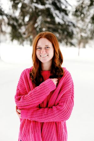 Person in bright pink sweater standing in snowy winter scene with evergreen trees.