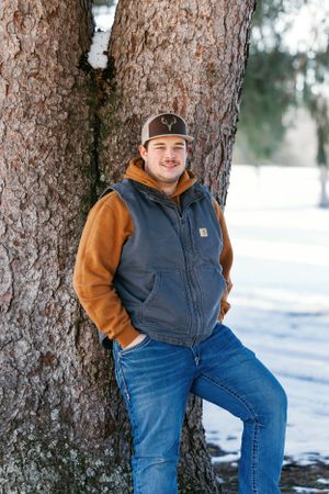 Person in workwear and baseball cap poses casually against large tree trunk in winter scenery.