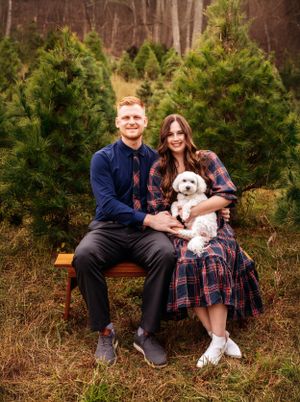 Couple sits on bench with small white dog among evergreen trees at Christmas tree farm.