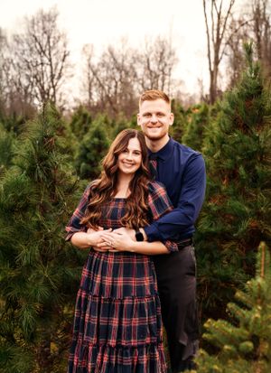 Couple embraces while posing for portrait among evergreen trees at Christmas tree farm.