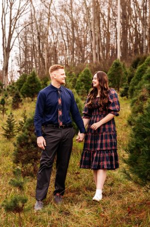 Couple in formal attire poses together at Christmas tree farm surrounded by evergreens.