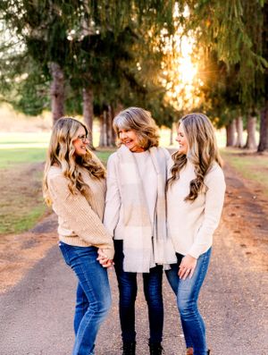Three people in neutral sweaters share a candid moment on tree-lined path at sunset.