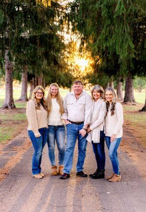 Family portrait of five people in matching cream sweaters and jeans standing on a path surrounded by evergreen trees.
