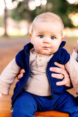 Baby wearing a navy blue vest and cream sweater sits while looking at camera with bright blue eyes.