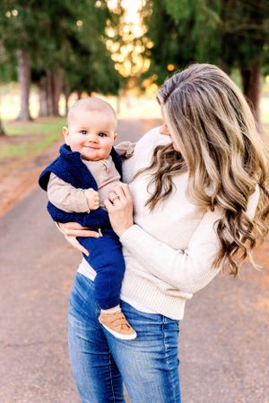 Baby in navy blue outfit smiles while being held by woman in cream sweater on outdoor path.