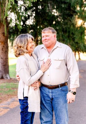 Couple in light colored outfits share a tender moment while standing on path with trees in background.
