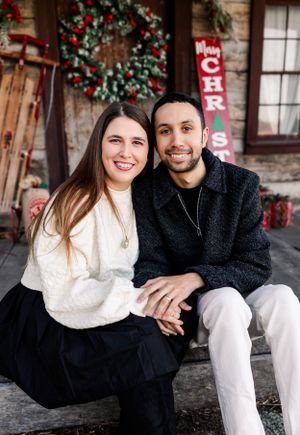 Couple in cozy winter sweaters sits on porch with Christmas decorations.
