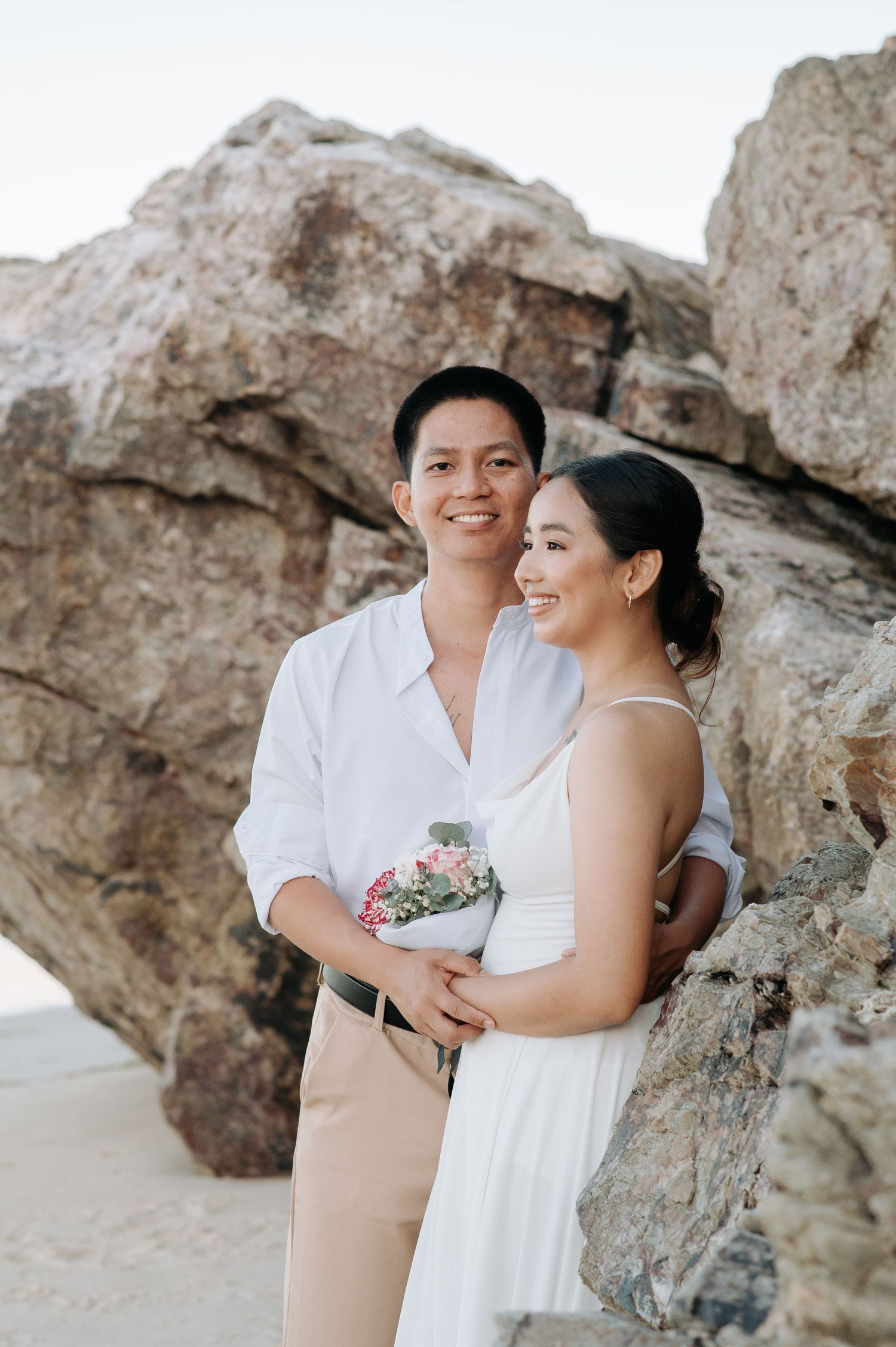 A couple poses with a small bouquet against large beach rocks during their engagement session.