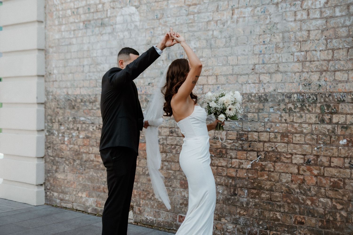 Couple performs elegant dance move against rustic brick wall with bridal bouquet.
