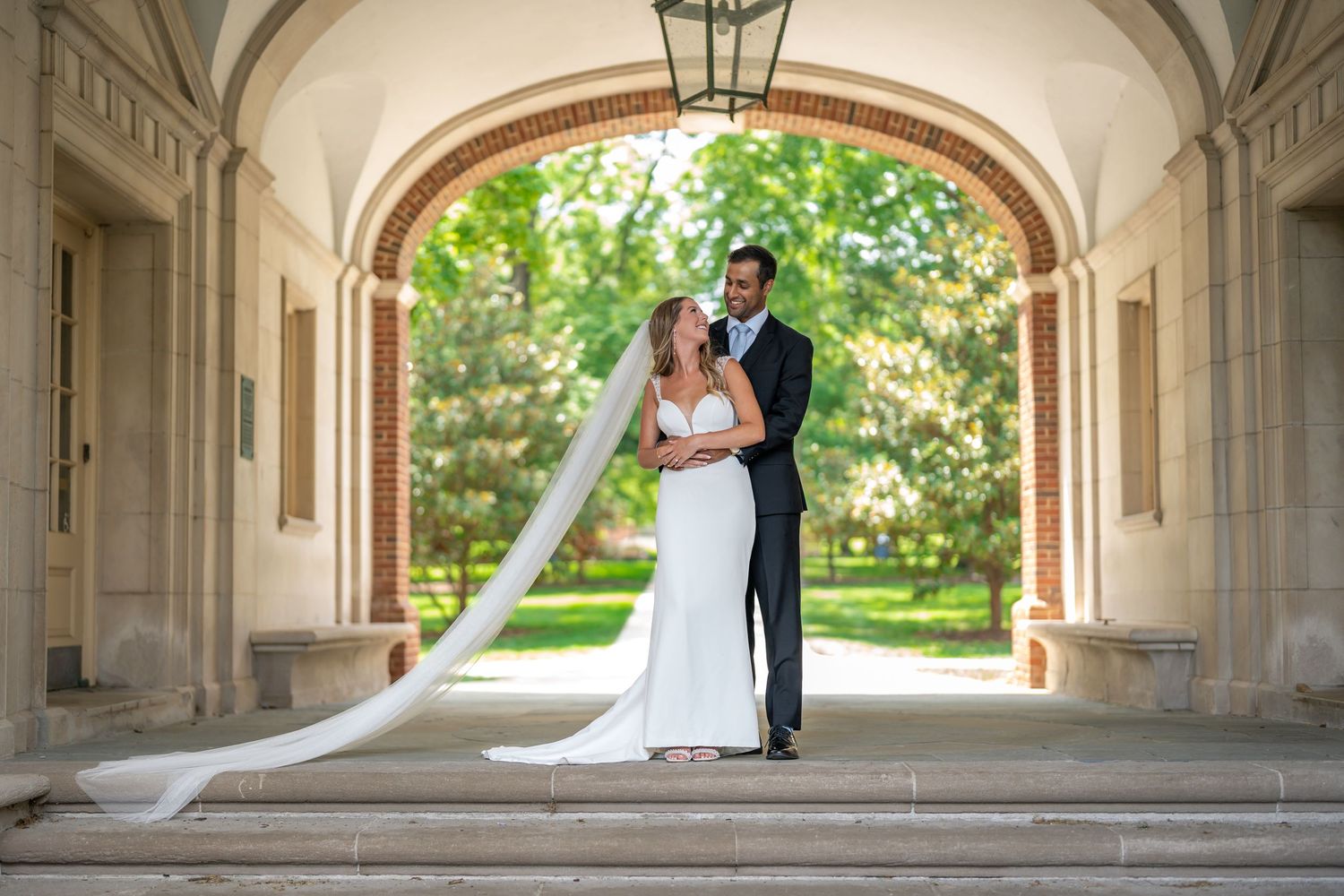 Wedding couple embracing under elegant stone archway with flowing veil.
