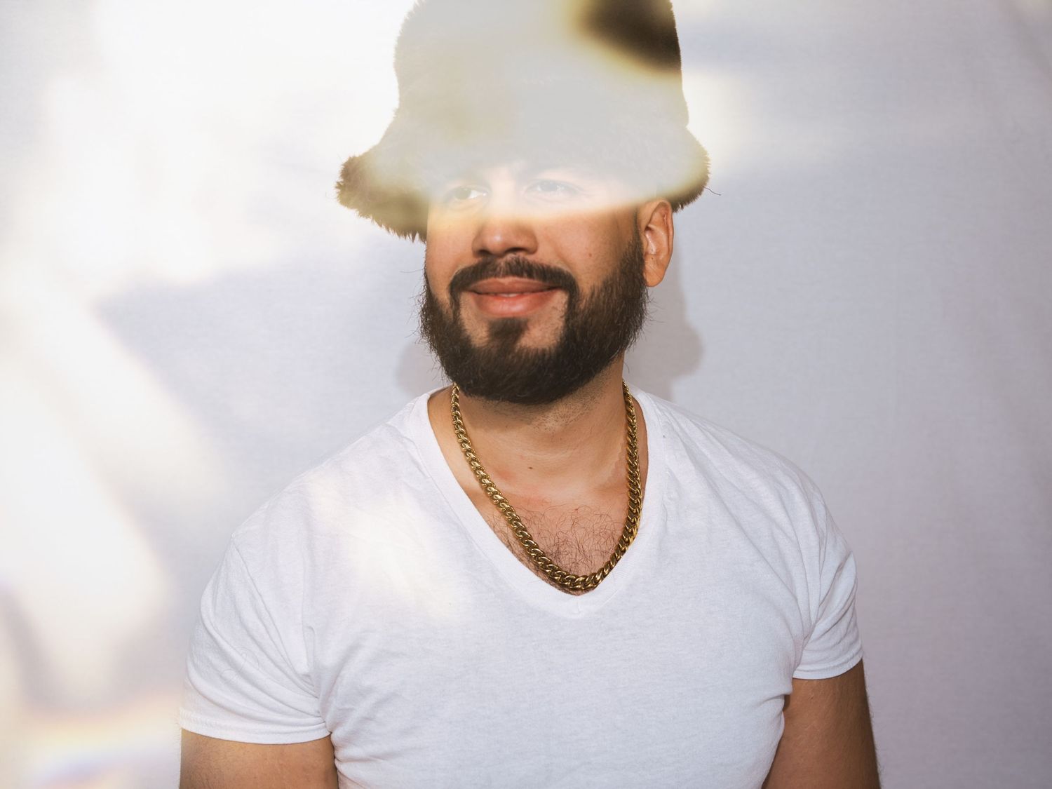 Man with beard and gold chain necklace wearing white t-shirt and hat, posing against light background.