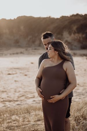 Pregnant woman and husband holding her belly on the beach.