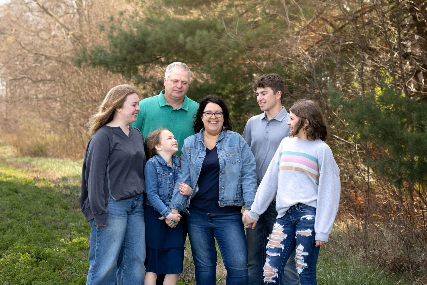 A family of 6 all smiling at mom next to a tree line.