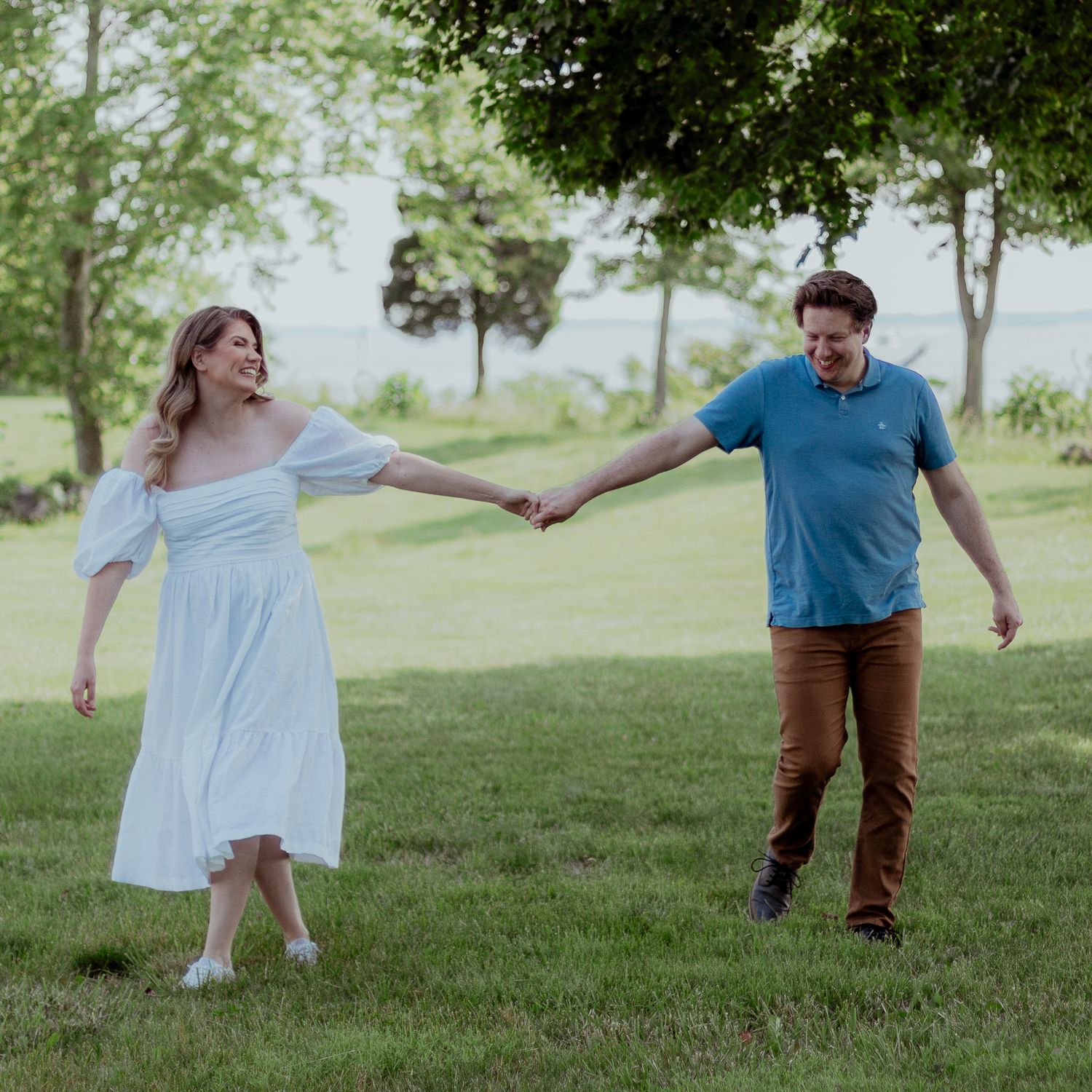 Couple holds hands while posing playfully in a sunlit grassy field.