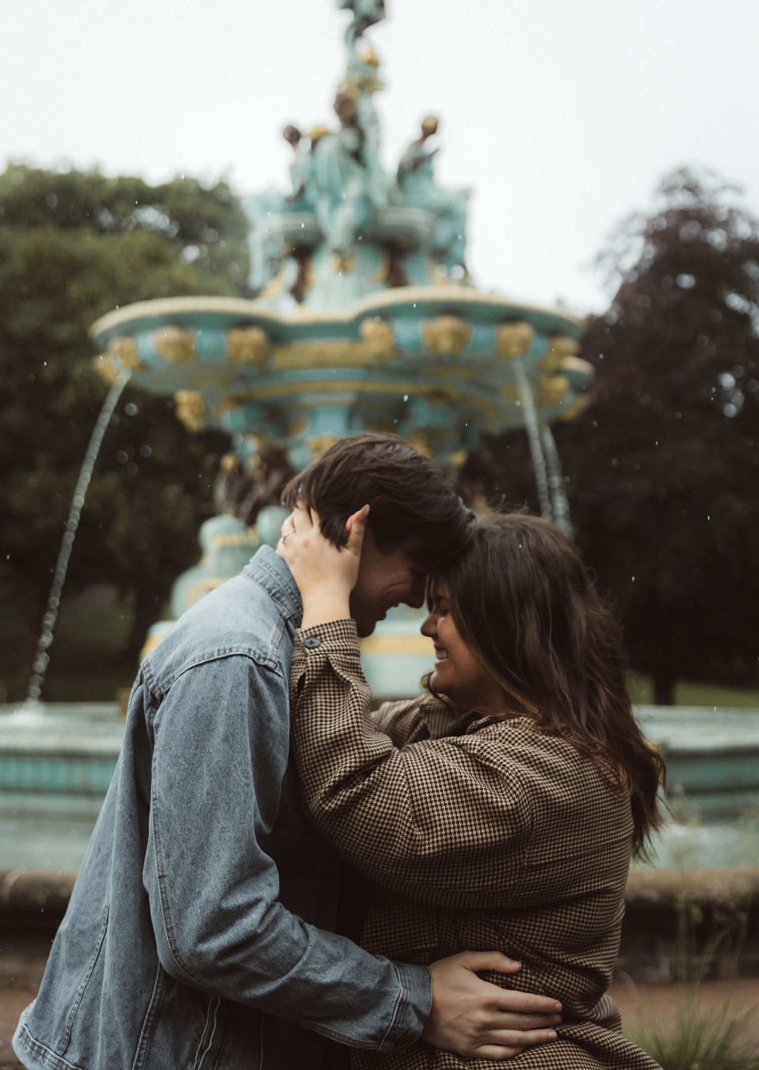 Two people share an intimate embrace near an ornate fountain in a park setting.