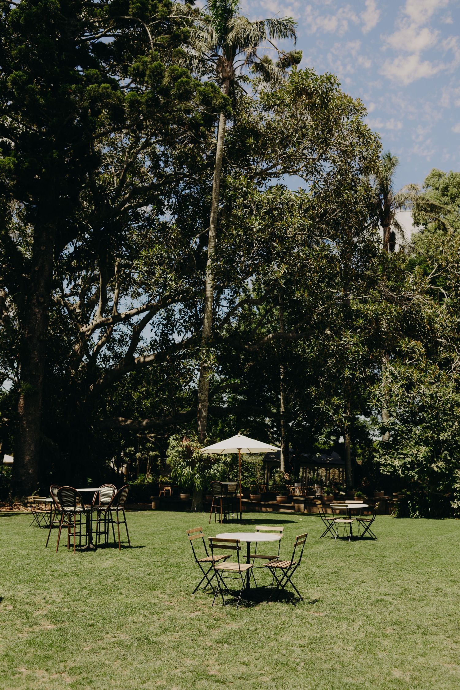 Outdoor wedding reception setup with scattered cocktail tables and chairs on a grassy lawn under trees.
