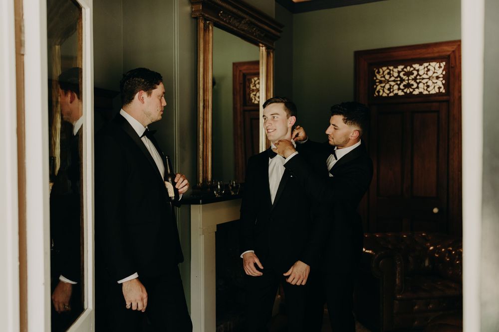 Groomsmen in black tuxedos gather in an elegant indoor venue for wedding photos.
