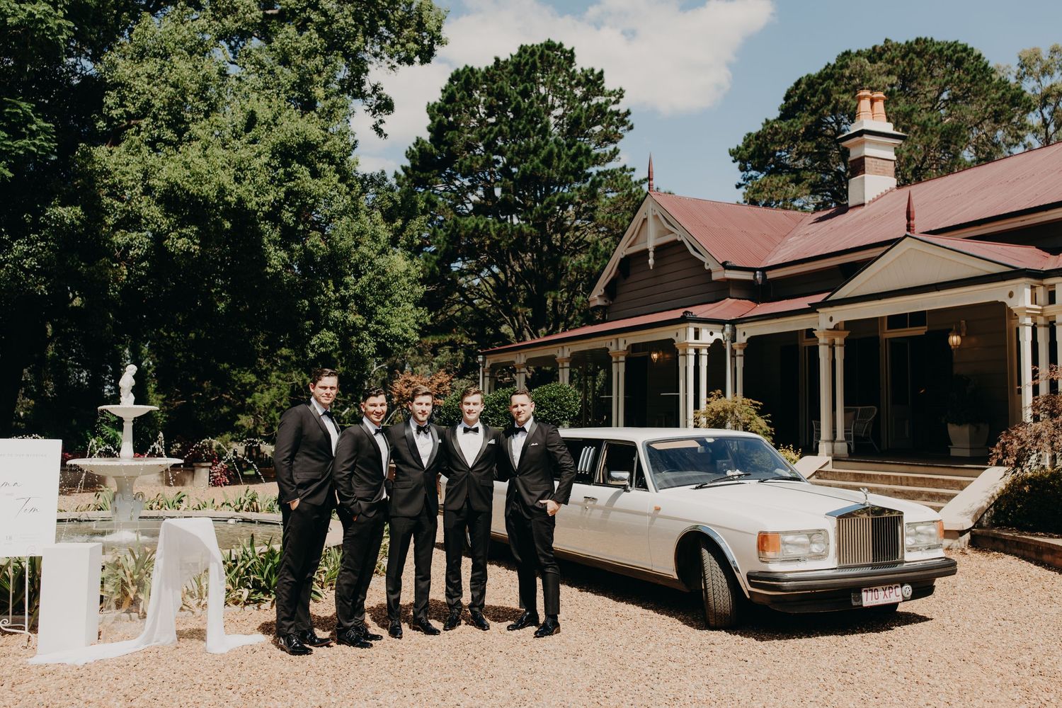 Wedding party and luxury car lined up outside a rustic venue on a sunny day.