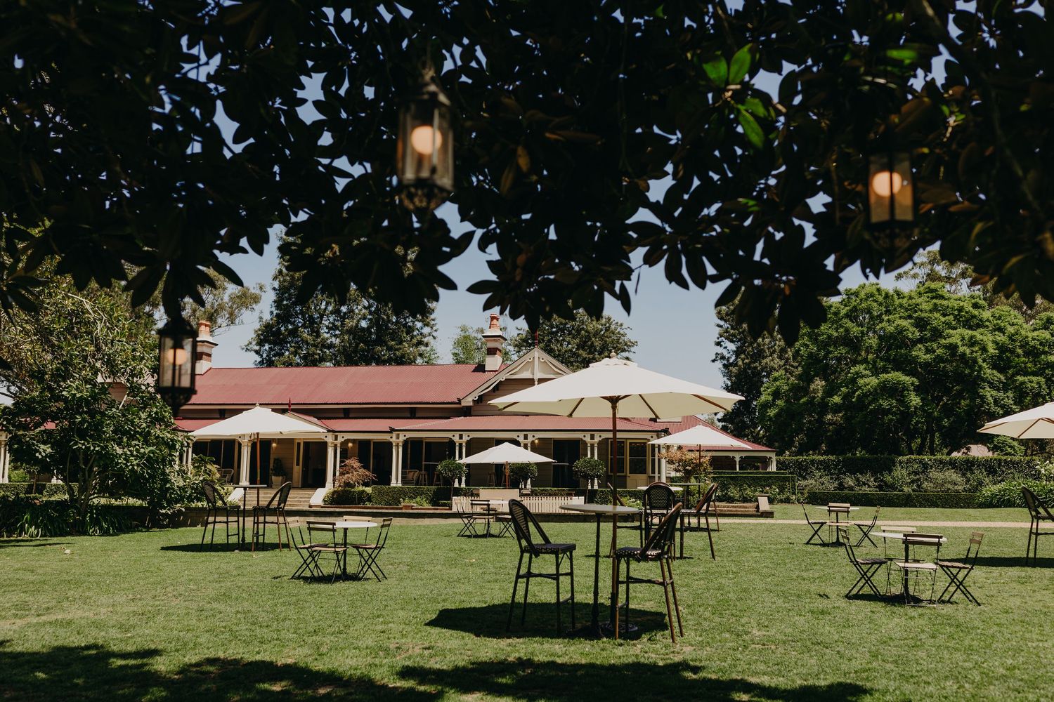 Outdoor wedding venue setup with bistro tables scattered across a green lawn near a colonial-style building.