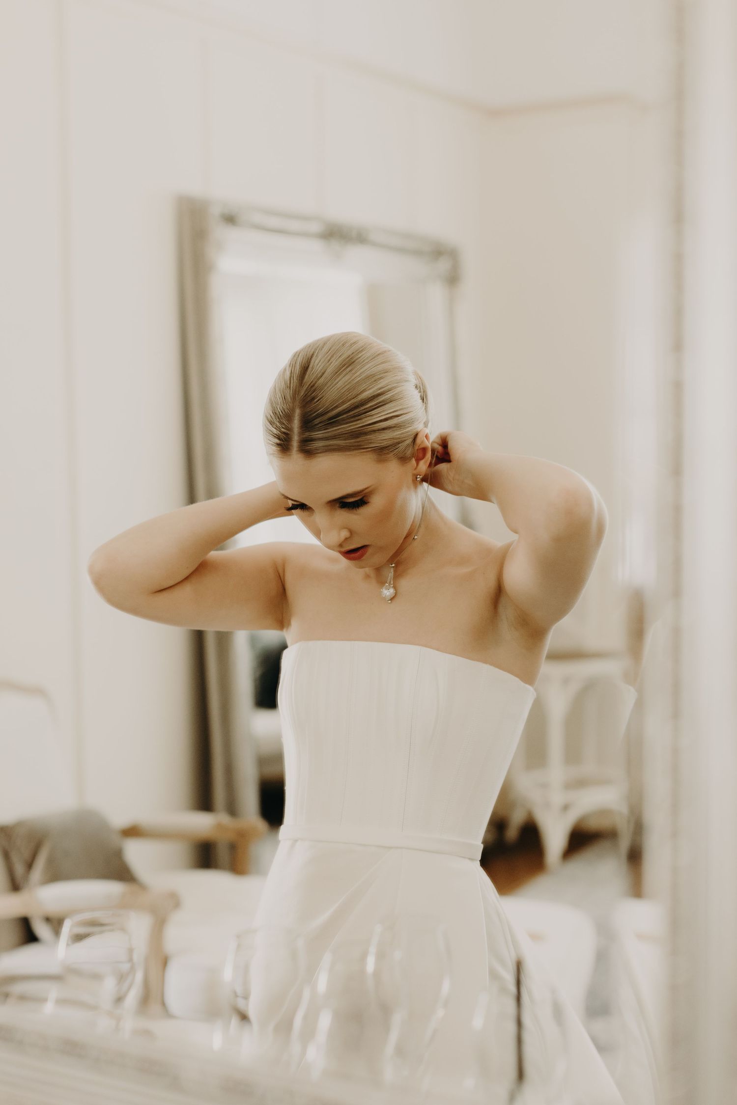 Bride in white strapless gown adjusting her hair in a bright room.
