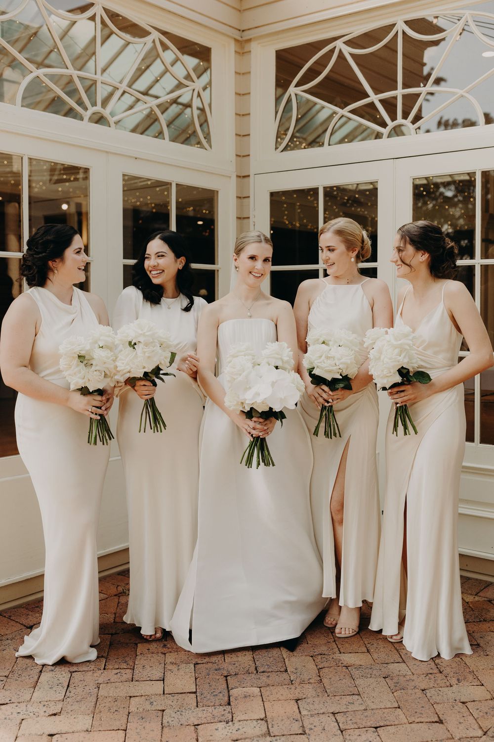 Bridesmaids in elegant white dresses holding bouquets pose in sunlit conservatory.