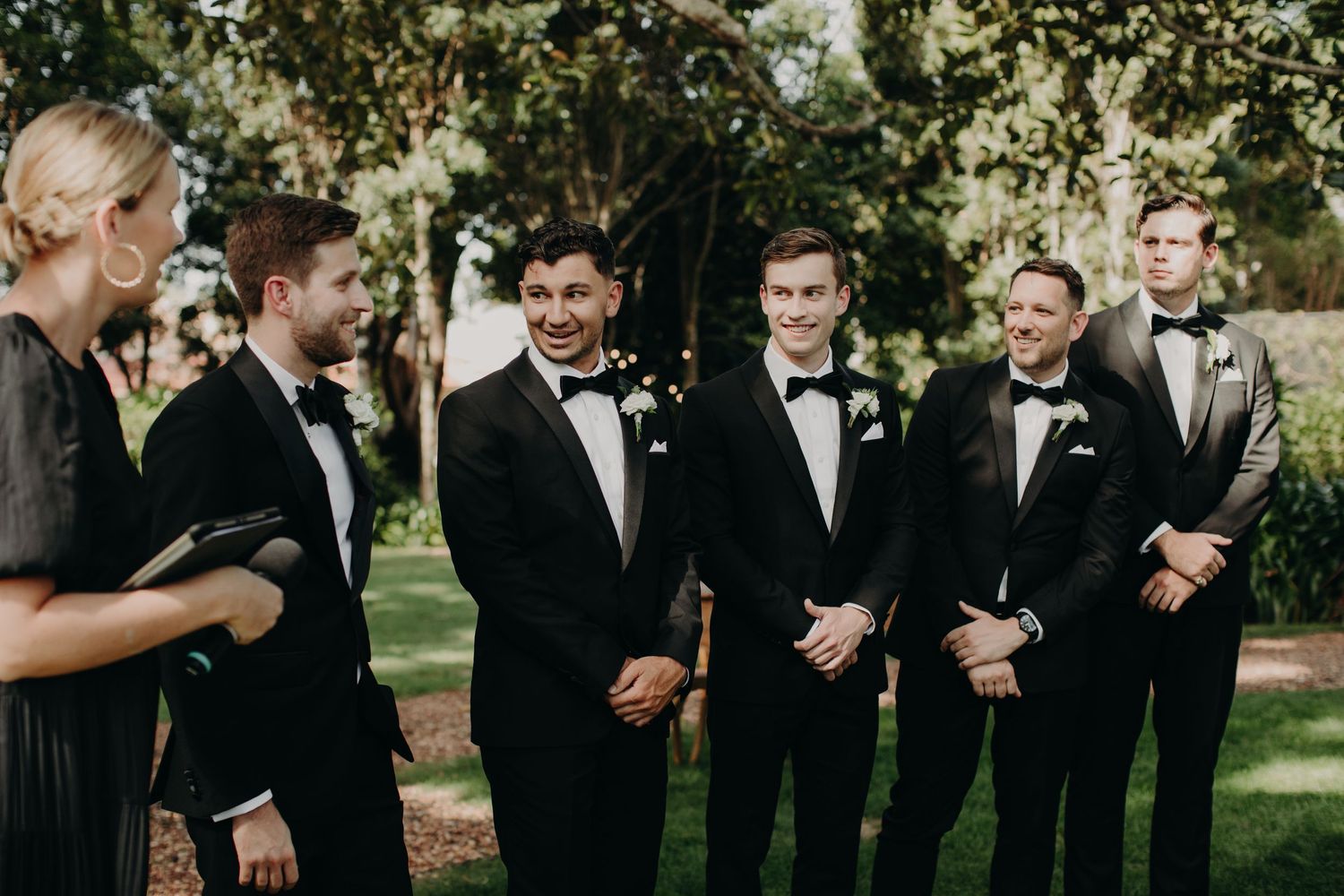 Groomsmen in black tuxedos laugh together during outdoor wedding photos.