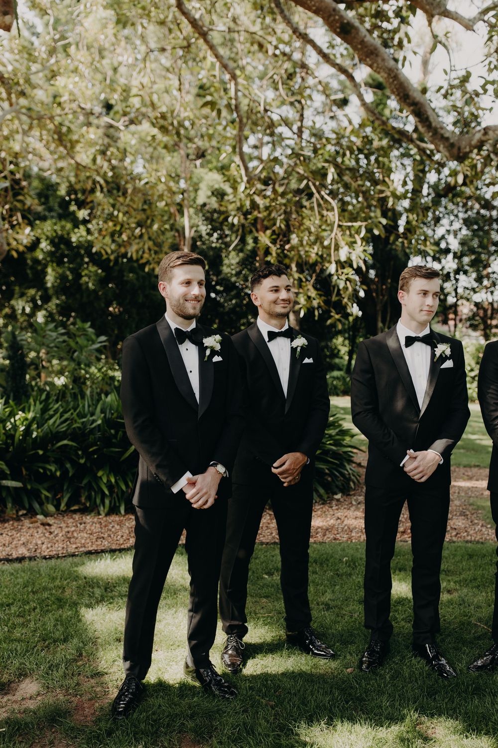 Groomsmen in black tuxedos stand in line against lush garden backdrop.