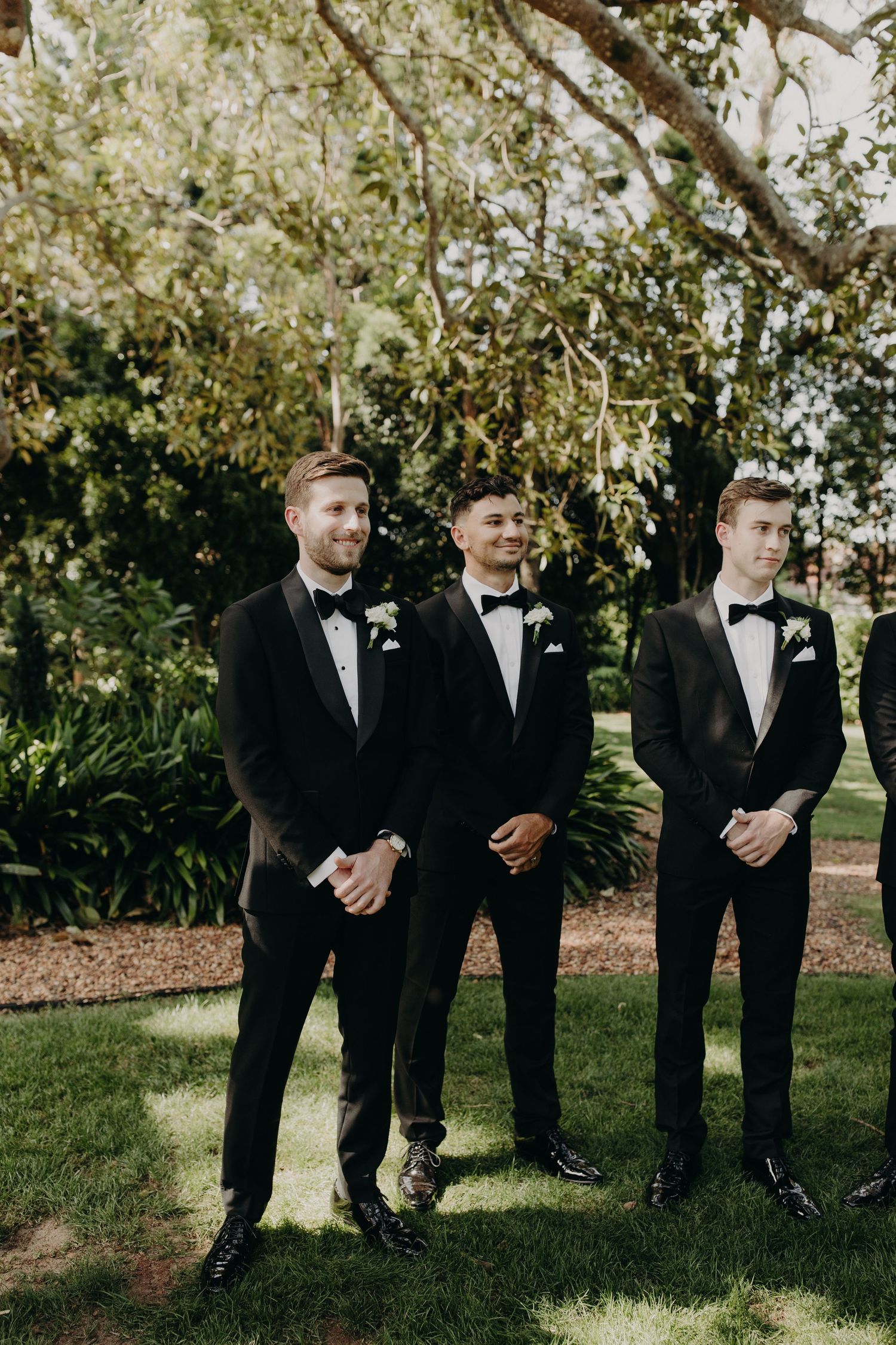 Groomsmen in black tuxedos stand in line against lush garden backdrop.