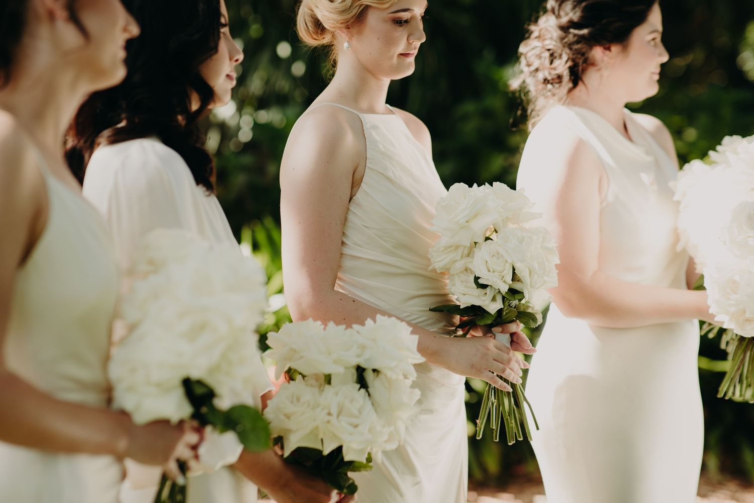 Bridesmaids in light colored dresses hold white floral bouquets during outdoor wedding.