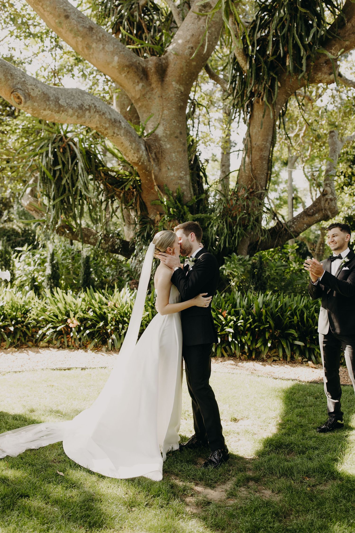 Wedding couple shares intimate moment under large tree during outdoor ceremony.