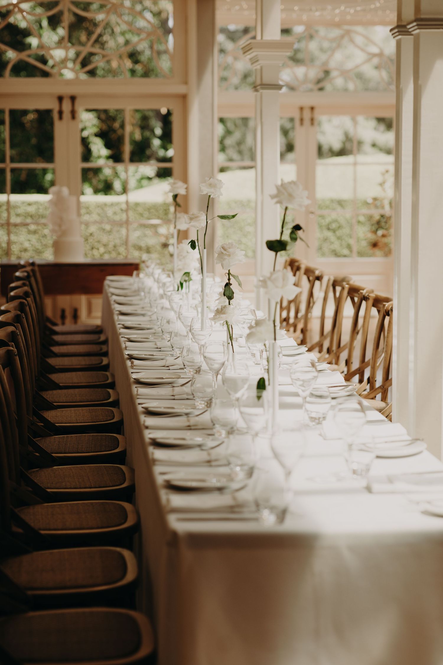 Elegant wedding reception table setting with crystal glasses and wooden chairs in a bright sunlit room.