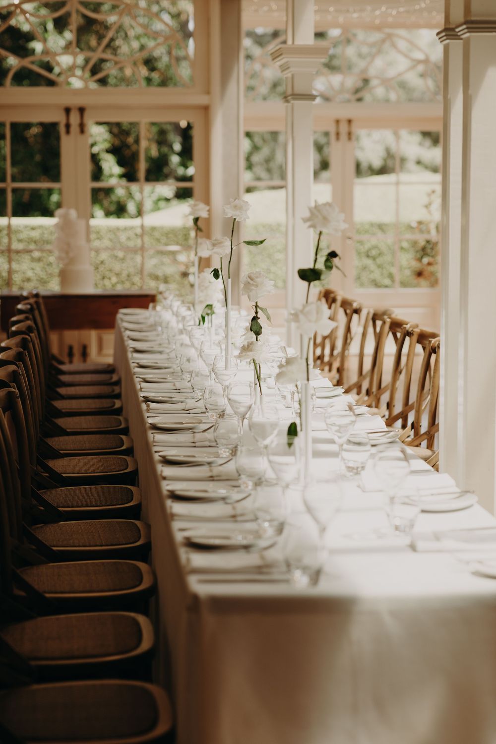 Elegant wedding reception table setting with crystal glasses and wooden chairs in a bright sunlit room.