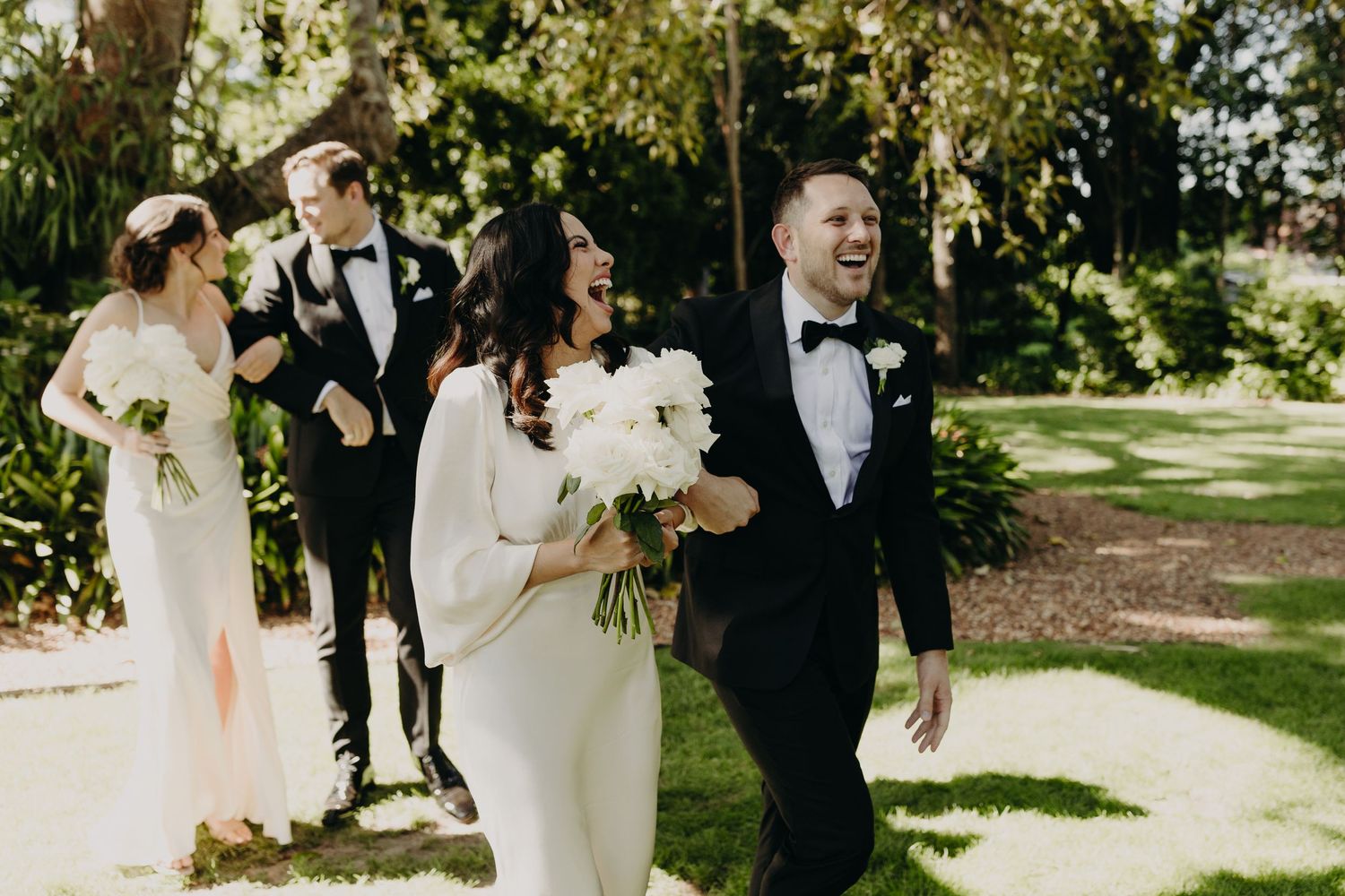 Wedding party poses together under trees during outdoor garden celebration.