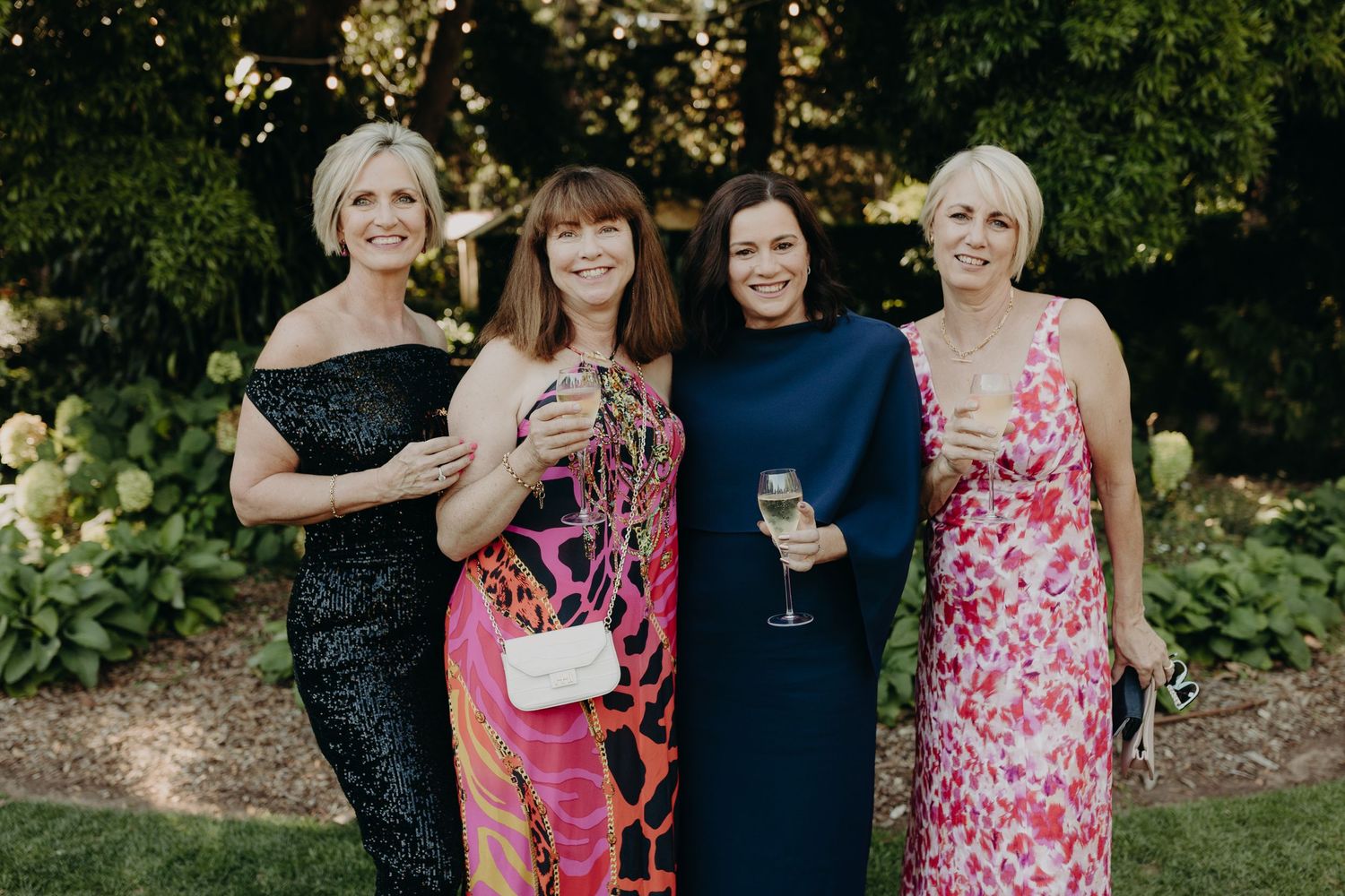 A group of wedding guests in colorful floral dresses gather outdoors for a photo.