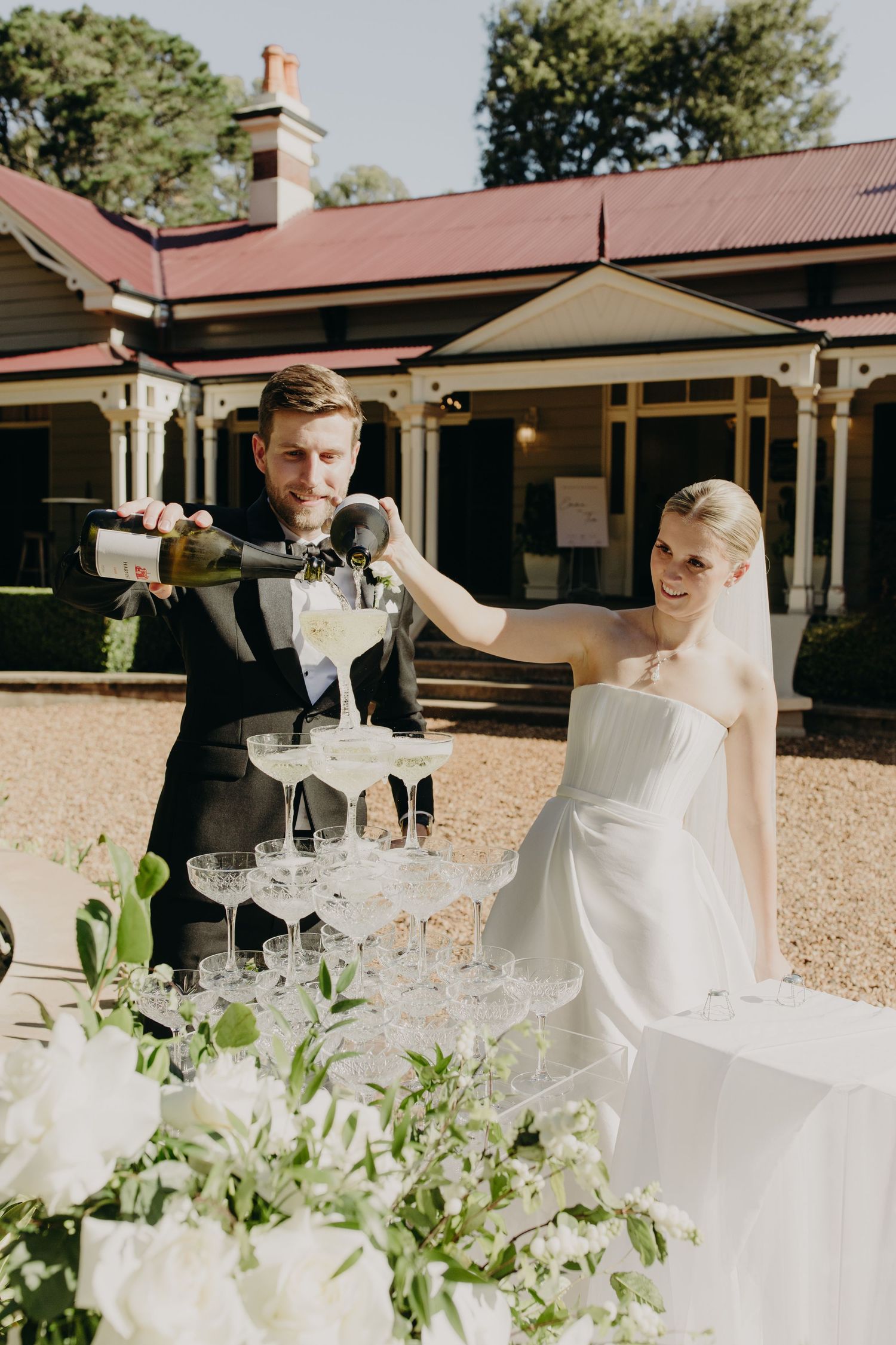 Wedding reception setup outdoors with elegant white table settings and greenery decorations.