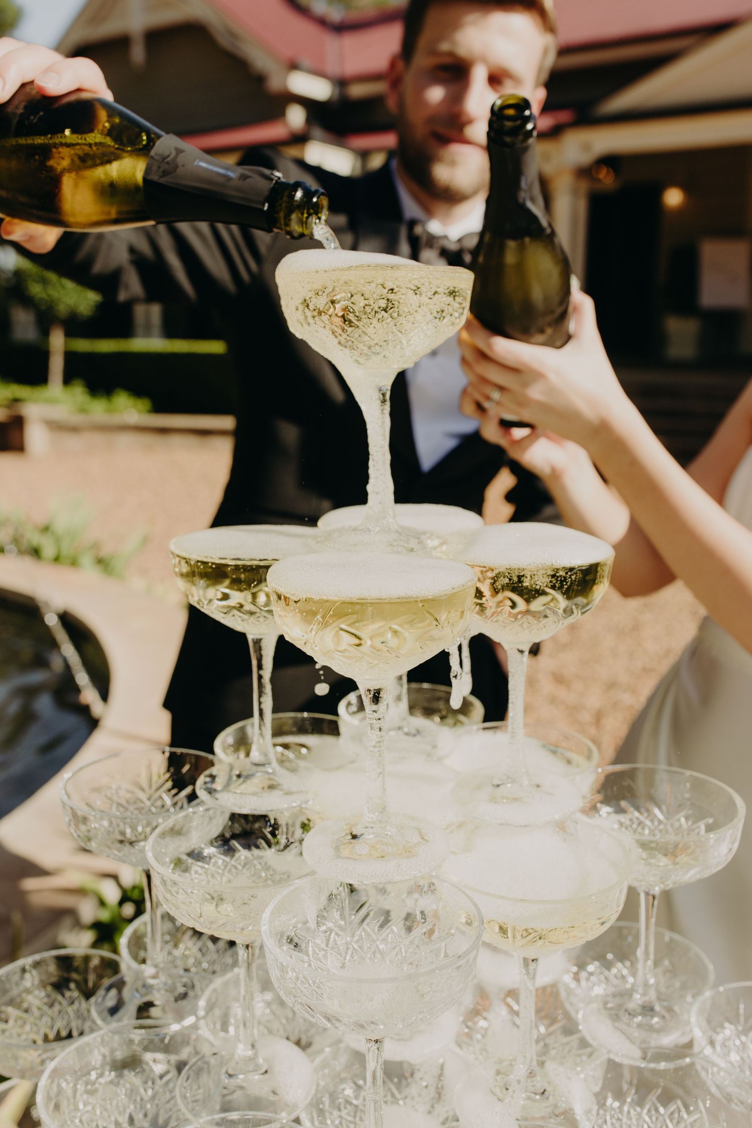 A champagne tower being filled with sparkling wine at an outdoor wedding celebration.