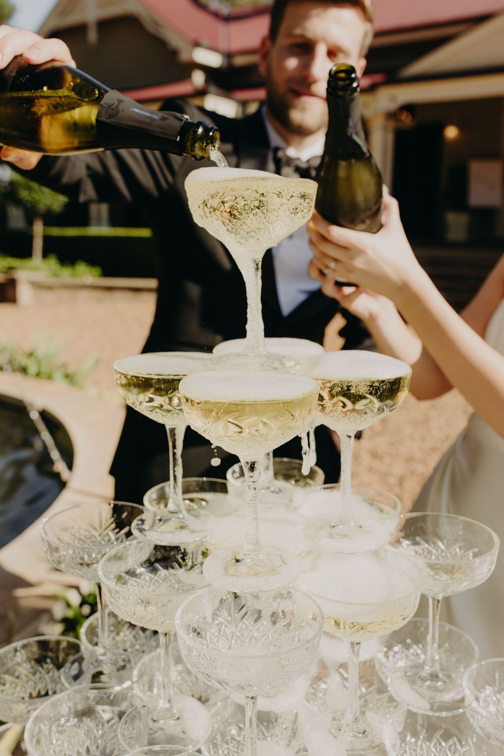 A champagne tower being filled with sparkling wine at an outdoor wedding celebration.