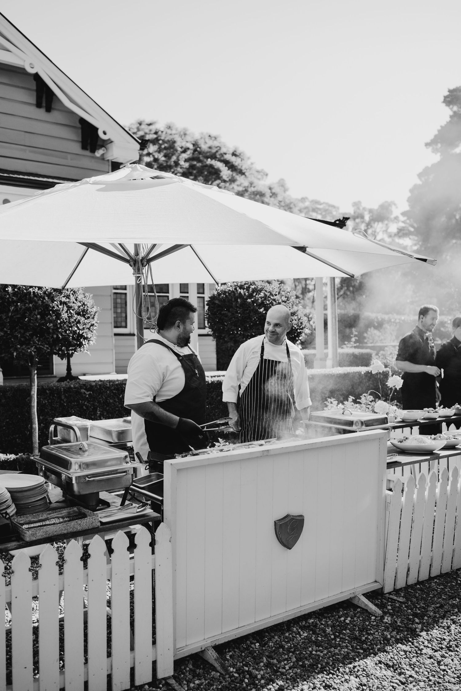 White picket fence lined outdoor catering station with chefs preparing food under market umbrellas.