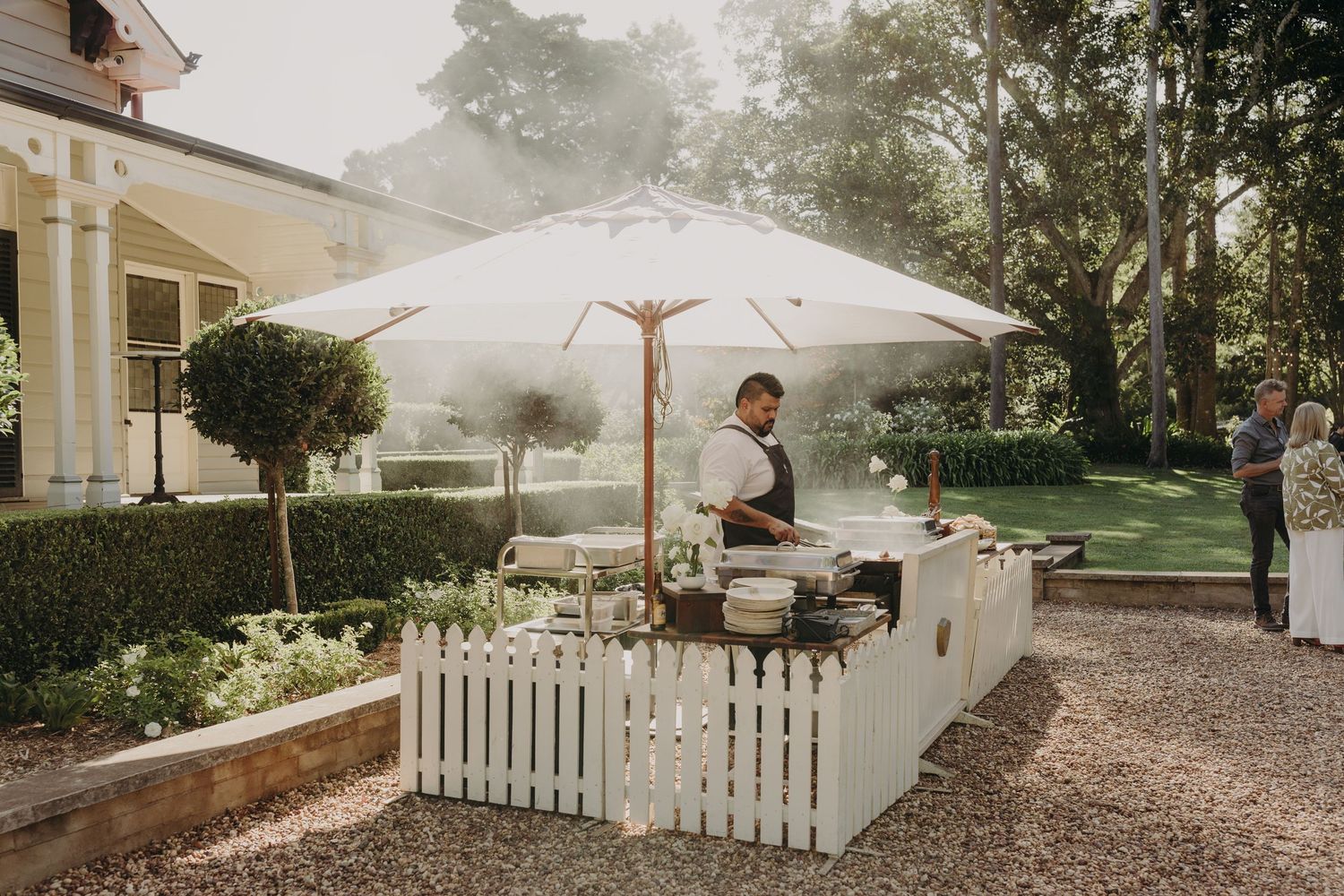 A serene outdoor venue with white picket fences and market umbrellas set up for an event.