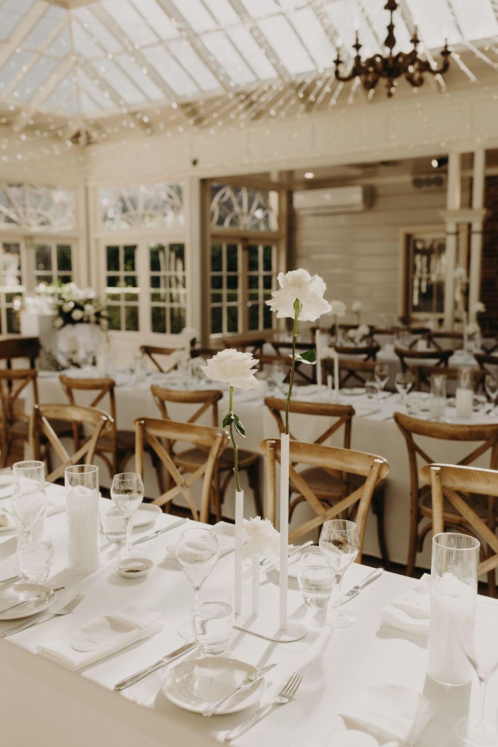 Elegant wedding reception tables set with white linens and rustic wooden chairs in a bright conservatory setting.