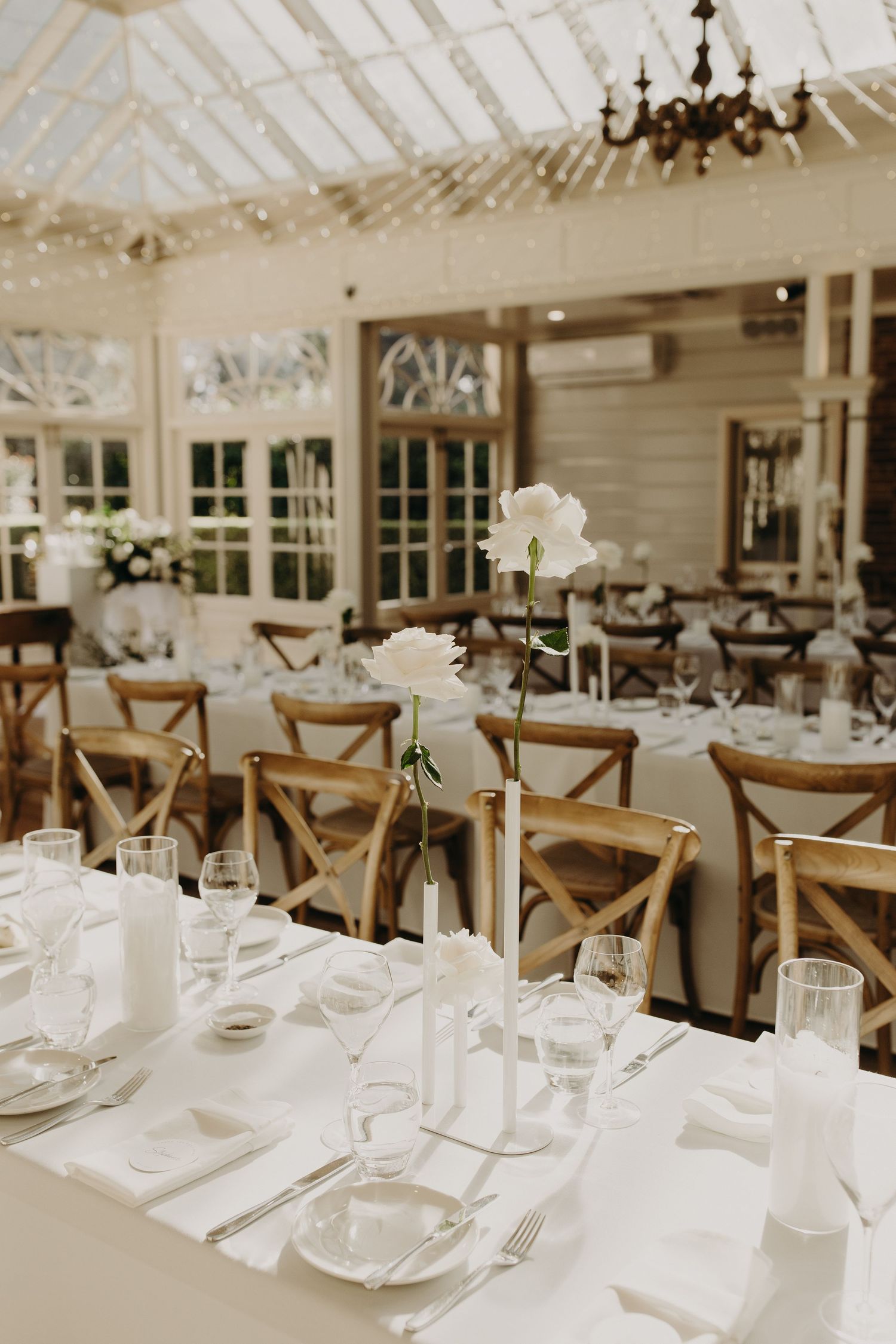 Elegant wedding reception tables set with white linens and rustic wooden chairs in a bright conservatory setting.