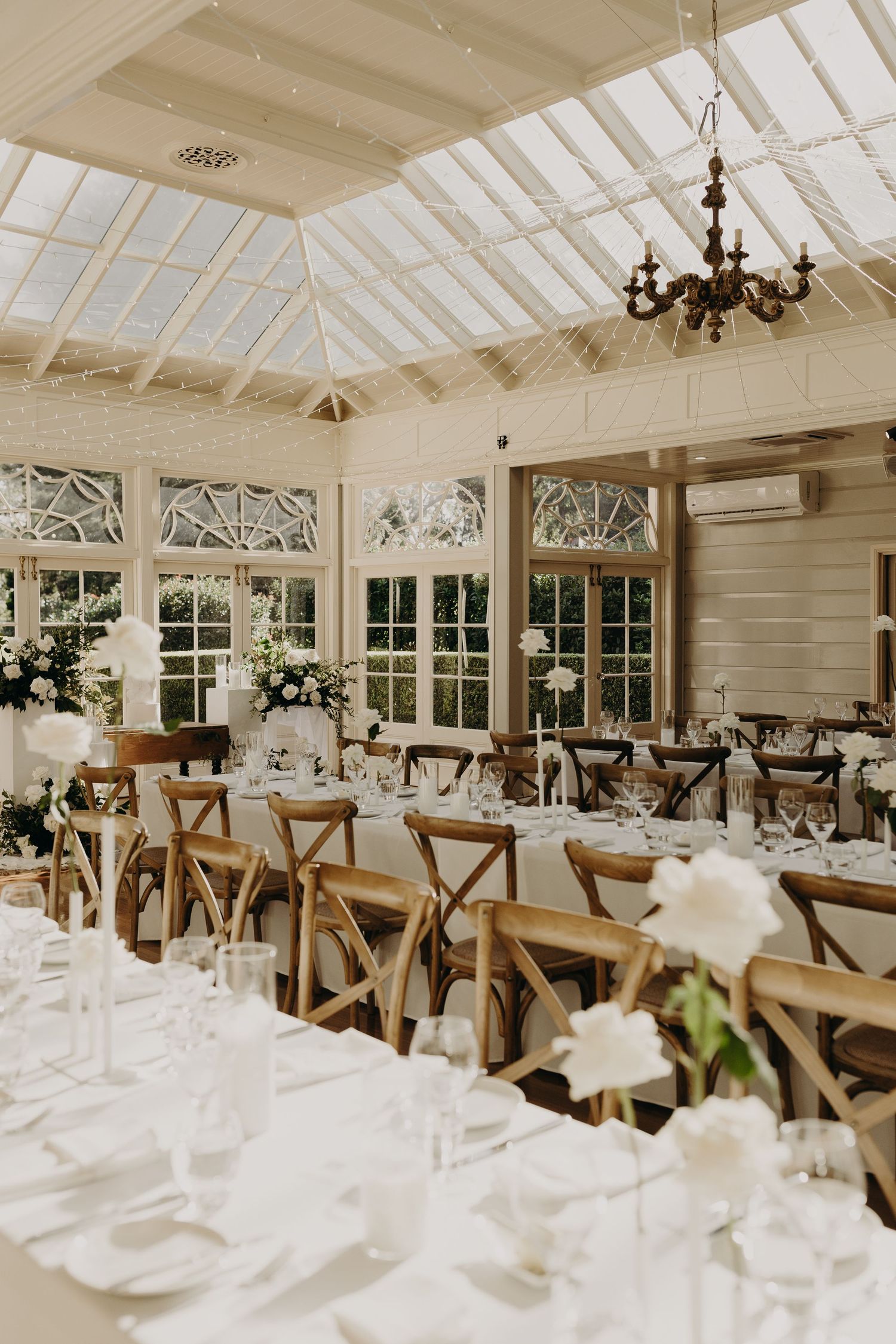 White-themed wedding reception venue with cross-back chairs and glass ceiling in natural light.