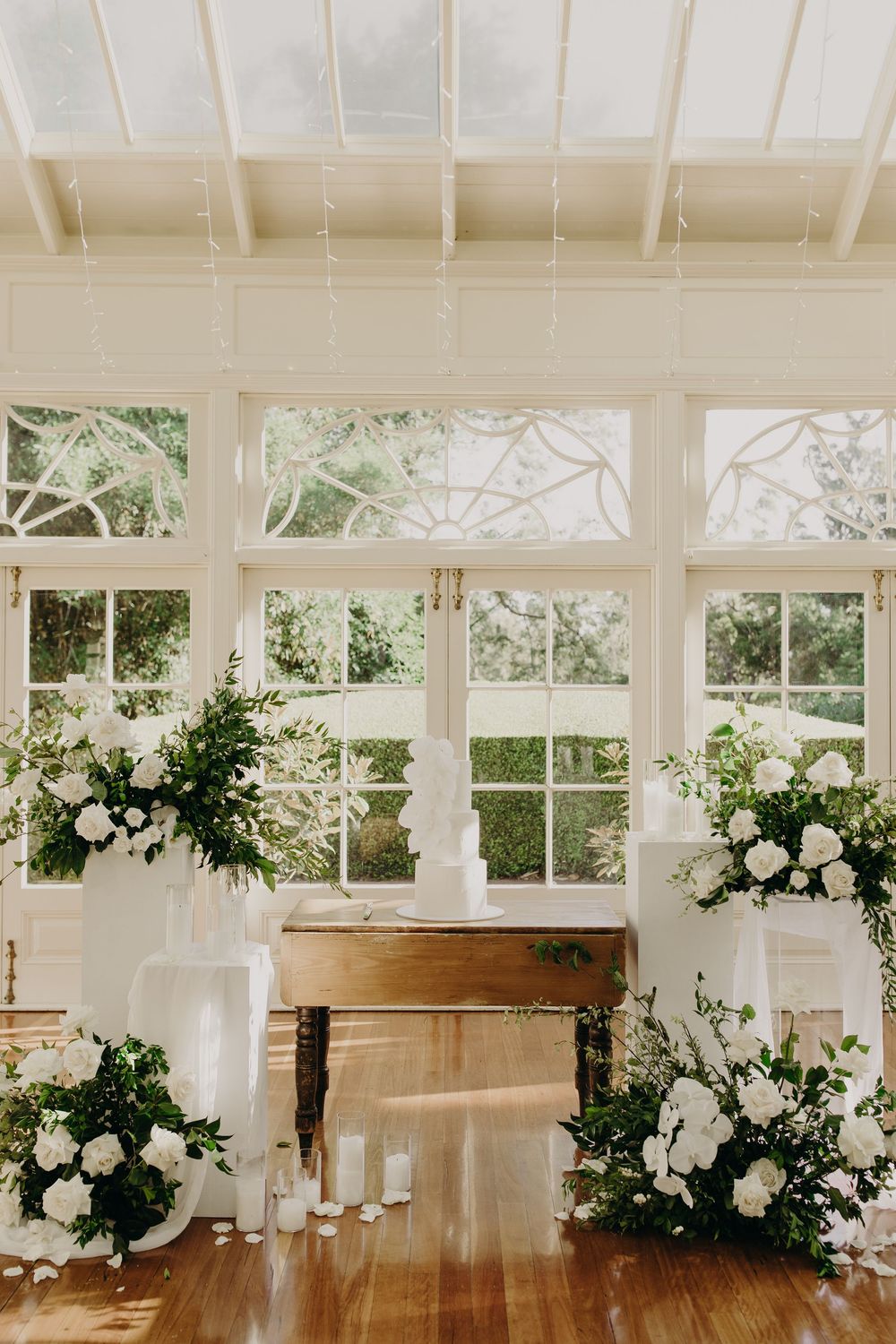 Sunlit conservatory decorated with white floral arrangements and greenery for an elegant wedding ceremony.