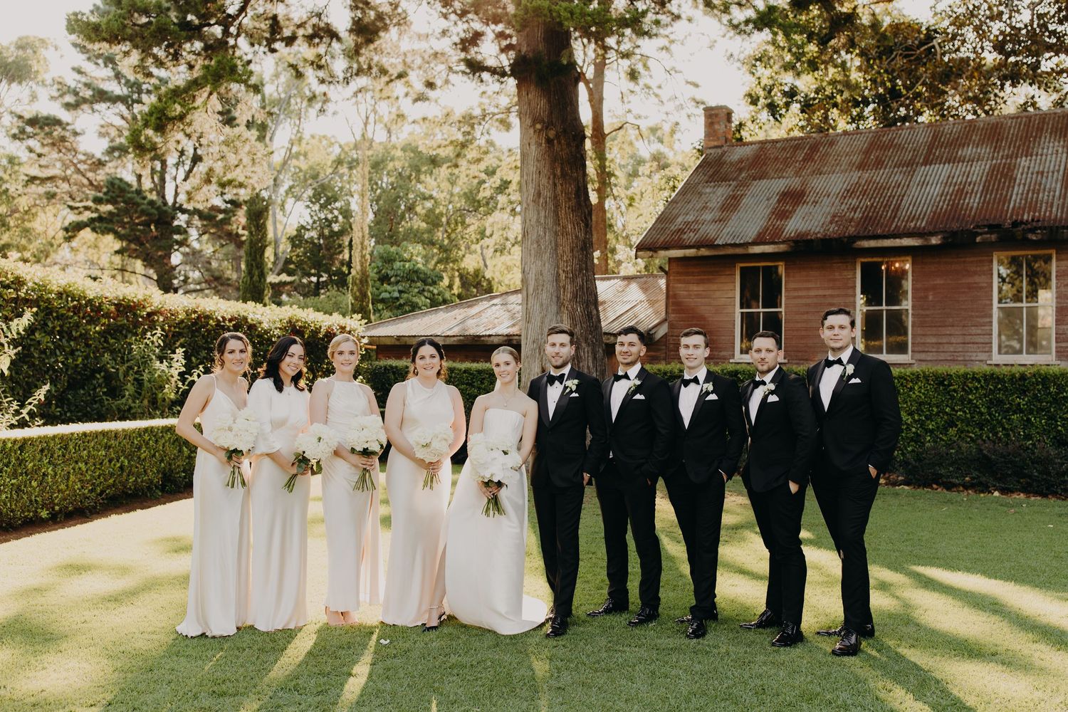 Wedding party in formal attire posed outdoors on manicured lawn with rustic building in background.