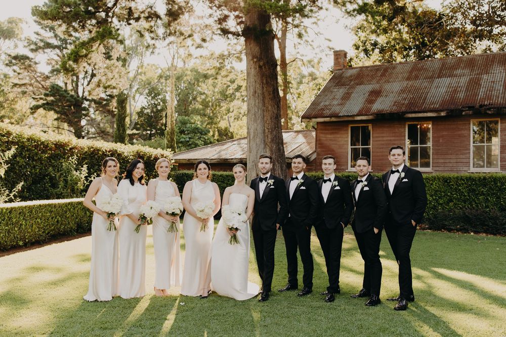 Wedding party in formal attire posed outdoors on manicured lawn with rustic building in background.