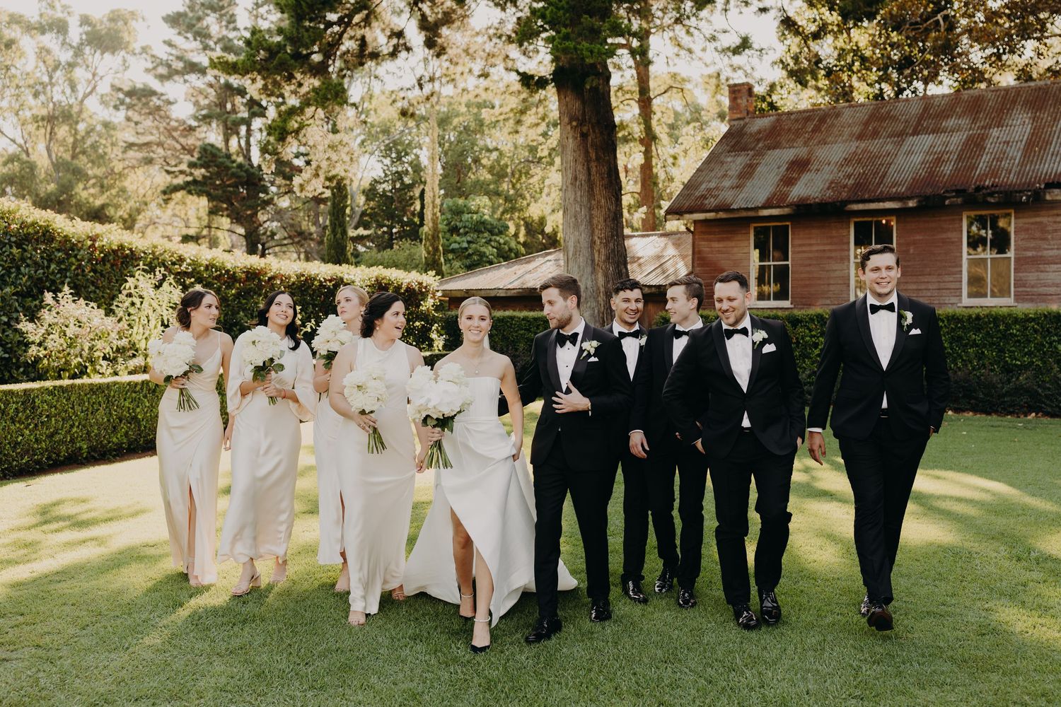 Wedding party in black and white attire standing on grass with rustic building in background.
