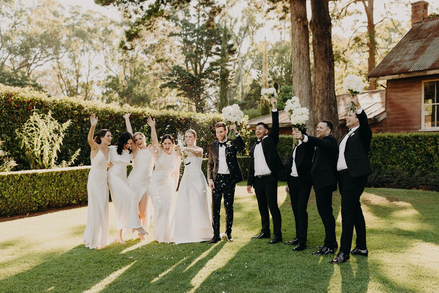 Wedding party celebrating outdoors with joyful poses against natural garden setting.