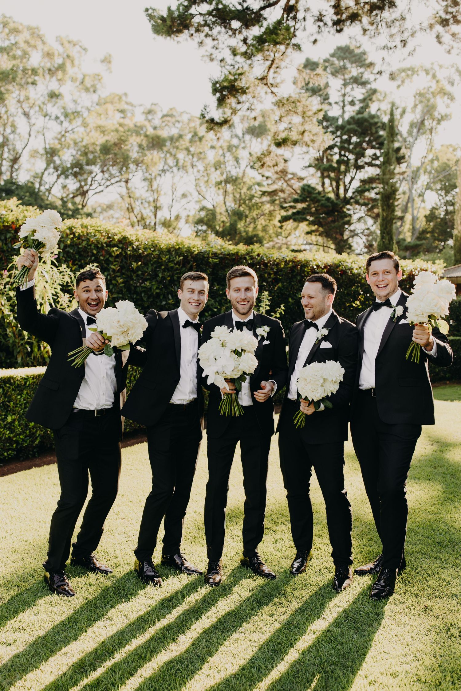 Groomsmen in black tuxedos pose with white bouquets on sunny lawn creating long shadows.